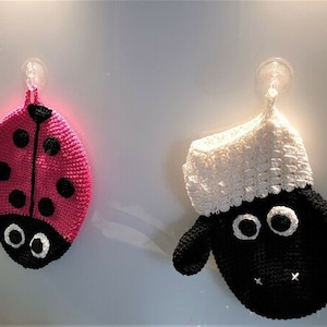 以下が含まれることがあります： ピンクの背景に黒い斑点のあるテントウムシの形をした、クロシェのキッチンタオル2枚。もう1枚は、白いふわふわの頭と黒い体をした羊の形をしたもの。