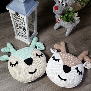 Könnte beinhalten: Zwei gehäkelte Rentier-Kissen, eines in hellgrün und weiß und das andere in braun und weiß. Beide Kissen haben gestickte Augen und eine schwarze Nase.