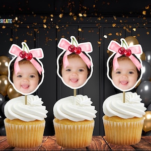 May include: Three cupcakes decorated with pink and white cherry toppers featuring a child's face. The cupcakes are on a black and gold background.