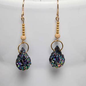 May include: A pair of gold-colored earrings with iridescent, dark blue, and purple druzy stone pendants. The earrings have gold-colored hooks, small gold beads, and a gold ring connecting the pendant. The earrings are displayed against a white background.