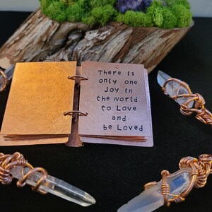 May include: A small copper-colored journal with the words "There is only one Joy in the world to Love and be Loved" is open. Clear quartz crystal pendants are wrapped in copper wire. A wooden base with moss and a purple crystal is in the background.