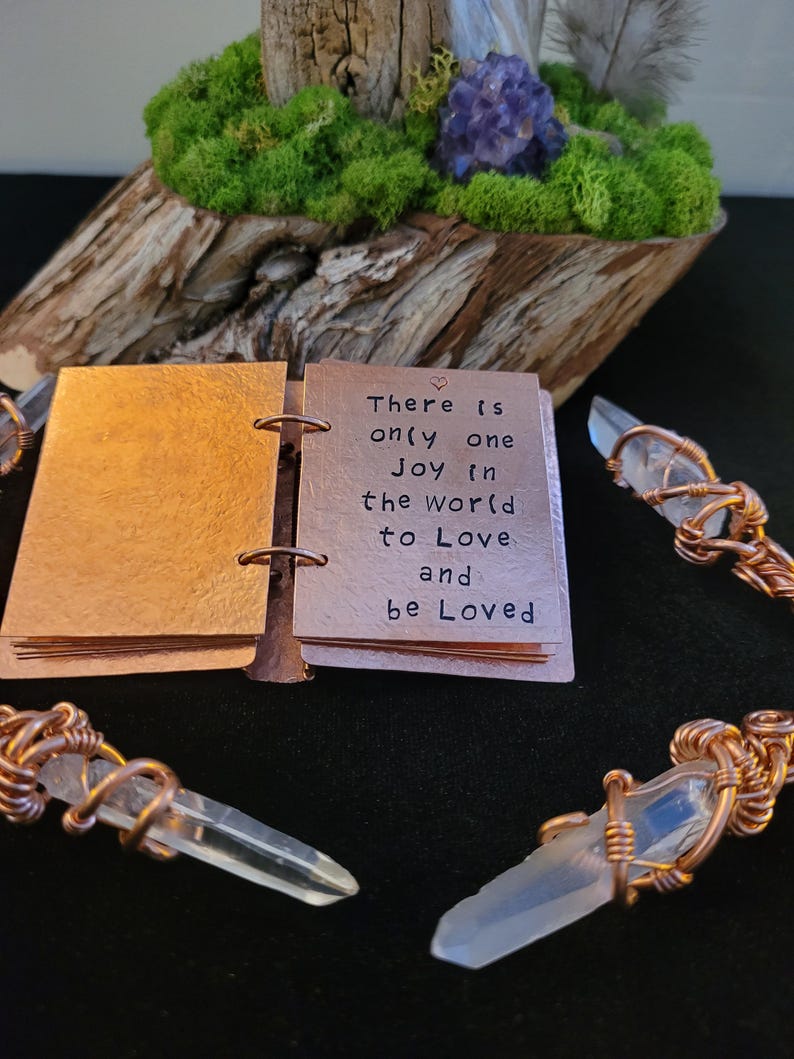 May include: A small copper-colored journal with the words "There is only one Joy in the world to Love and be Loved" is open. Clear quartz crystal pendants are wrapped in copper wire. A wooden base with moss and a purple crystal is in the background.