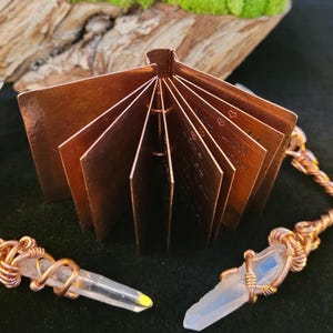 May include: A small, open copper book with etched pages, displayed with two crystal pendants wrapped in copper wire. The book is positioned in front of a dark background, with a wooden and mossy element in the background.
