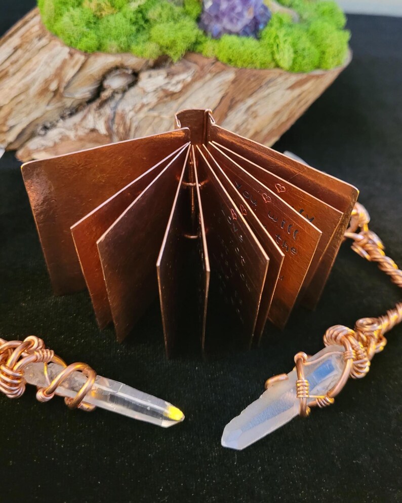 May include: A small, open copper book with etched pages, displayed with two crystal pendants wrapped in copper wire. The book is positioned in front of a dark background, with a wooden and mossy element in the background.