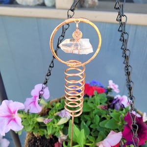 May include: A copper-colored hanging plant stake with a clear quartz crystal and a coiled spring design. The stake is suspended by a black chain and is positioned above a hanging basket filled with pink, red, and purple petunias.