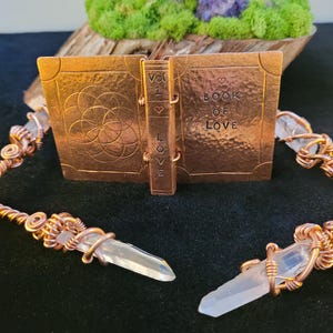 May include: A copper-colored metal book sculpture with the words "BOOK OF LOVE" and "VOL 1 LOVE" etched on the cover and spine. Two clear quartz crystal pendants are connected to the book with copper wire. The background includes moss, wood, and amethyst.