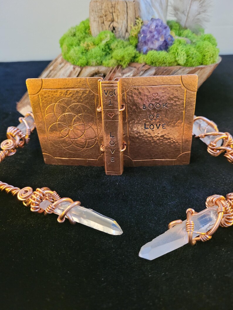 May include: A copper-colored metal book sculpture with the words "BOOK OF LOVE" and "VOL 1 LOVE" etched on the cover and spine. Two clear quartz crystal pendants are connected to the book with copper wire. The background includes moss, wood, and amethyst.