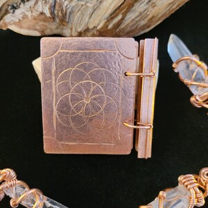 May include: A small, copper-colored book with a geometric design on the cover. The book is open, revealing the pages. Clear quartz crystals are wrapped in copper wire, creating a mystical aesthetic.