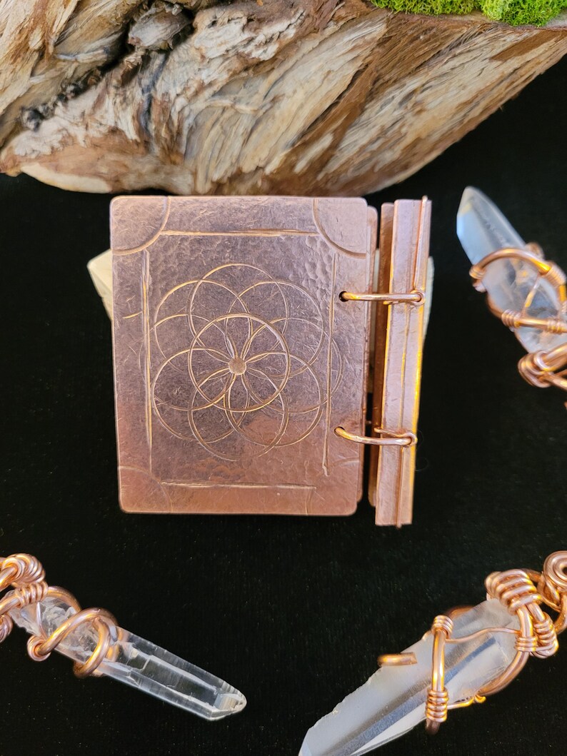 May include: A small, copper-colored book with a geometric design on the cover. The book is open, revealing the pages. Clear quartz crystals are wrapped in copper wire, creating a mystical aesthetic.