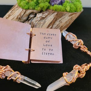 May include: A small copper-colored notebook with the quote "The first duty of Love is to listen" is open. Clear quartz crystals are wrapped in copper wire. The background includes a wooden base with green moss, a purple crystal, and feathers.