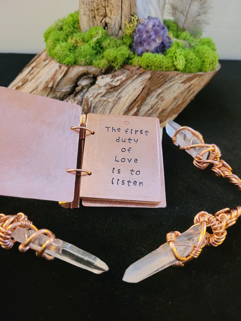 May include: A small copper-colored notebook with the quote "The first duty of Love is to listen" is open. Clear quartz crystals are wrapped in copper wire. The background includes a wooden base with green moss, a purple crystal, and feathers.