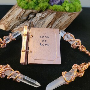 May include: A small, copper-colored book with the words "BOOK OF LOVE" stamped on the cover, accented with a heart symbol. The book is surrounded by clear quartz crystals wrapped in copper wire. The background features a wooden base with green moss and a purple crystal.