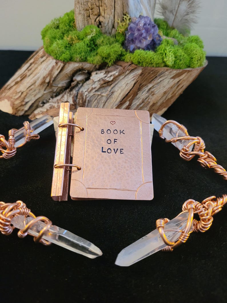 May include: A small, copper-colored book with the words "BOOK OF LOVE" stamped on the cover, accented with a heart symbol. The book is surrounded by clear quartz crystals wrapped in copper wire. The background features a wooden base with green moss and a purple crystal.