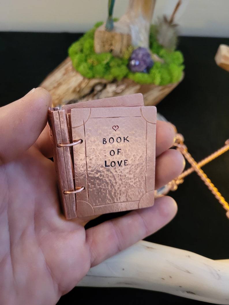May include: A small, copper-colored book with the words "BOOK OF LOVE" and a heart symbol etched on the cover. The miniature book is held in a hand, showcasing its intricate details and craftsmanship. The book's pages are visible, and it is bound with a metal ring.