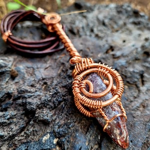 May include: A pendant featuring a pointed, mottled brown and cream stone encased in intricate copper wire wrapping. The wire forms a spiral cage around the stone and extends upward into a bail, attached to a dark brown leather cord necklace.