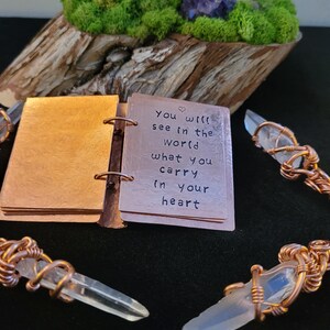 May include: A small, open copper-colored book with the quote "You will see in the world what you carry in your heart" is displayed. Clear quartz crystals are wrapped in copper wire. The background includes a wooden base with green moss and a purple flower.