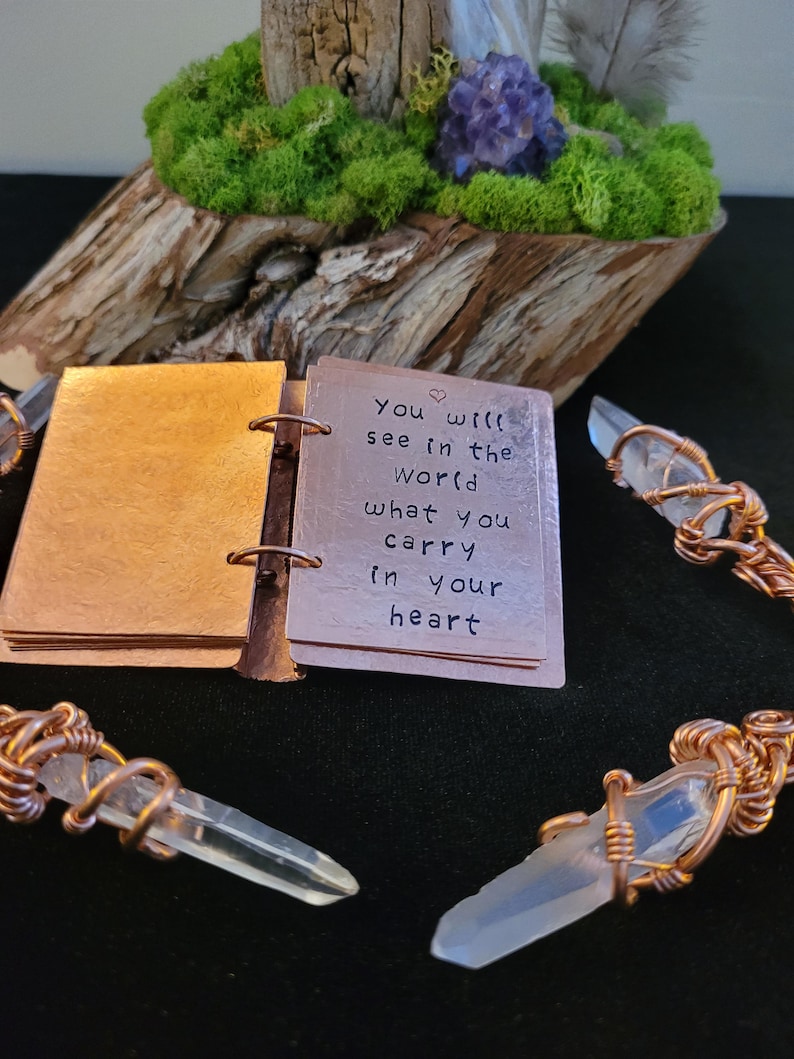 May include: A small, open copper-colored book with the quote "You will see in the world what you carry in your heart" is displayed. Clear quartz crystals are wrapped in copper wire. The background includes a wooden base with green moss and a purple flower.