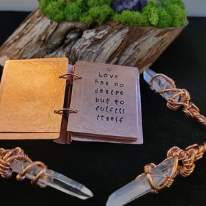 May include: A copper-colored journal with the quote "Love has no desire but to fulfill itself" is open, next to two crystal pendants with copper wire wrapping. The background features a wooden base with moss and a purple crystal.