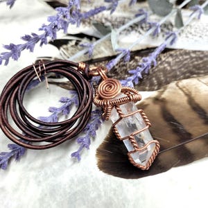 May include: A copper wire-wrapped crystal pendant necklace with a brown leather cord. The clear crystal is encased in a spiral and twisted wire design. The necklace is displayed with purple lavender sprigs and a feather.