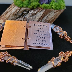 May include: A small copper-colored journal with the quote "To change the world we must first change ourselves" is open. Clear quartz crystal points are wrapped in copper wire. A wooden base with moss and purple flowers is in the background.