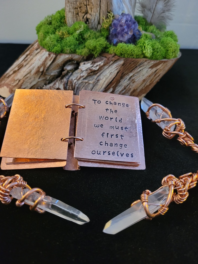 May include: A small copper-colored journal with the quote "To change the world we must first change ourselves" is open. Clear quartz crystal points are wrapped in copper wire. A wooden base with moss and purple flowers is in the background.