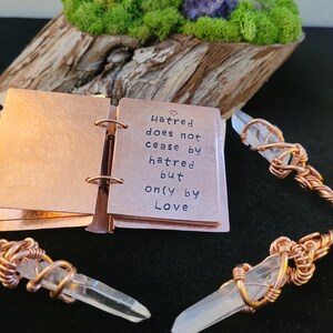 May include: A copper-colored mini-book with the quote "Hatred does not cease by hatred but only by Love." is open, next to two crystal pendants with copper wire wrapping. The background includes a wooden base with moss and a purple crystal.