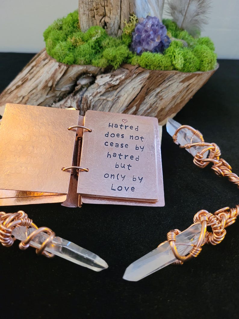 May include: A copper-colored mini-book with the quote "Hatred does not cease by hatred but only by Love." is open, next to two crystal pendants with copper wire wrapping. The background includes a wooden base with moss and a purple crystal.