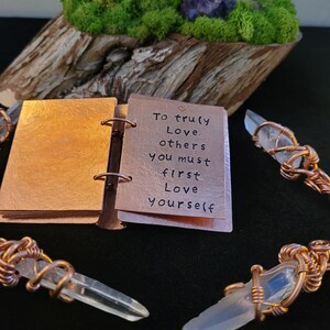 May include: A copper-colored mini-book with the quote "To truly love others you must first love yourself." is open, revealing the message. Clear quartz crystals are wrapped in copper wire. A wooden base with moss and a purple crystal cluster is in the background.