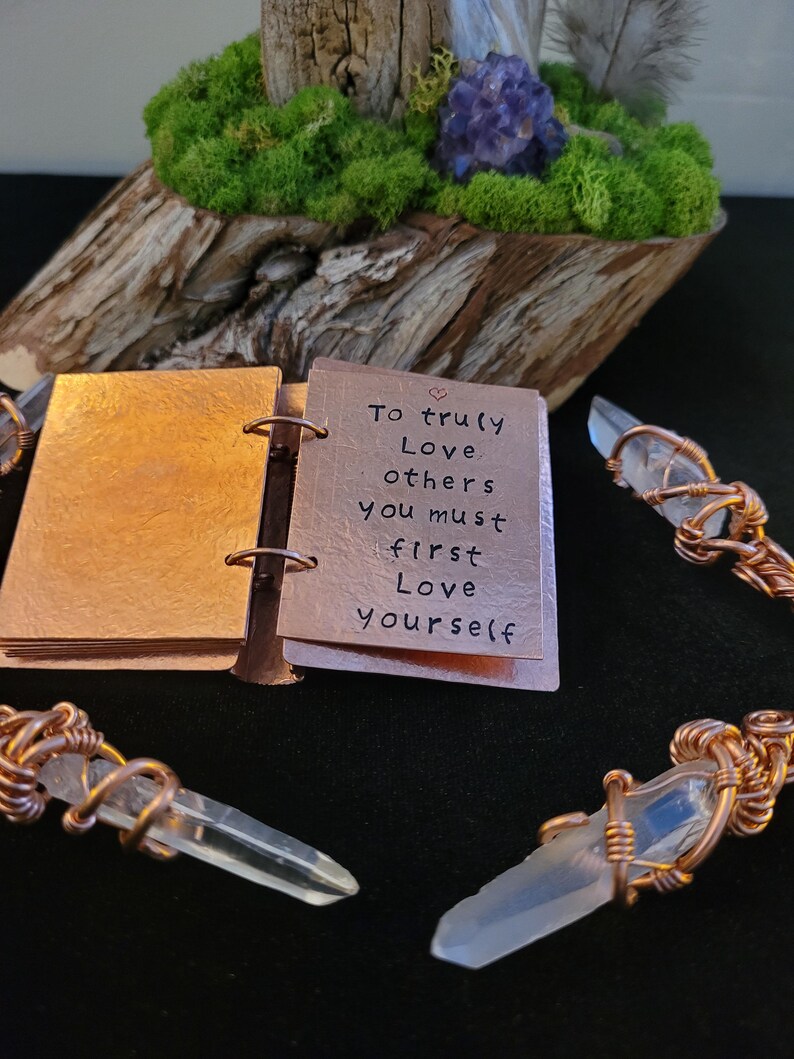 May include: A copper-colored mini-book with the quote "To truly love others you must first love yourself." is open, revealing the message. Clear quartz crystals are wrapped in copper wire. A wooden base with moss and a purple crystal cluster is in the background.