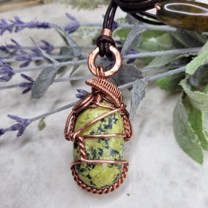 May include: A handmade pendant necklace featuring a green and black stone wrapped in copper wire. The stone is suspended from a copper ring and brown leather cord. The necklace is a unique piece of jewelry.