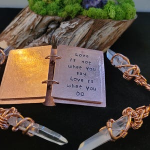 May include: A copper-colored mini-book with the quote "Love is not what you say, Love is what you DO" is open, next to wire-wrapped quartz crystal pendants. The background includes a wooden base with moss and a purple crystal.