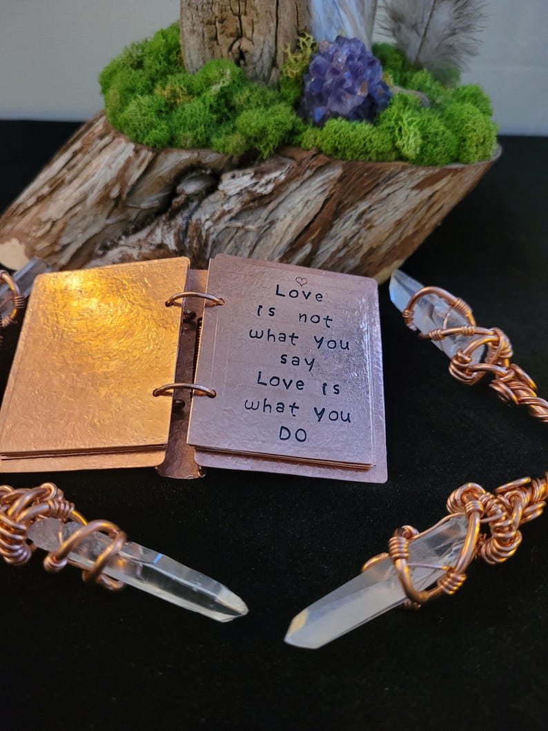 May include: A copper-colored mini-book with the quote "Love is not what you say, Love is what you DO" is open, next to wire-wrapped quartz crystal pendants. The background includes a wooden base with moss and a purple crystal.