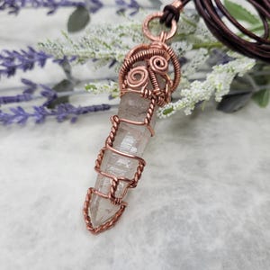 May include: A clear quartz crystal pendant wrapped in copper wire. The wire is intricately woven around the crystal, forming decorative patterns. The pendant hangs from a brown leather cord. The background includes lavender and white flowers.