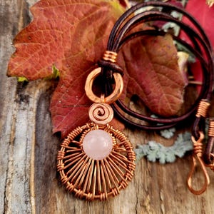 May include: A handcrafted copper pendant necklace featuring a rose quartz gemstone. The pendant has a circular design with radiating wires and a spiral accent. The necklace has a brown leather cord and a copper clasp. The background includes fall foliage.