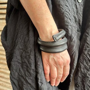 May include: A black leather bracelet with a unique twisted design. The bracelet is made of multiple layers of leather that are woven together to create a textured look.