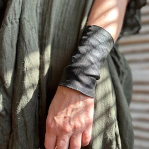 May include: A wide black leather cuff bracelet with a smooth, matte finish and a slightly wrinkled texture. The cuff wraps around the wrist, featuring clean, straight edges at the top and bottom, providing a simple and solid black appearance.
