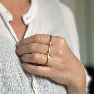 May include: A gold-colored ring with a vertical bar design. The ring is worn on a finger, and the image shows a close-up view of the hand and ring. The ring is a simple, modern piece of jewelry.