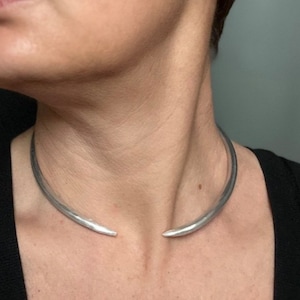 Puede incluir: Un collar choker plateado con un diseño simple y curvo.