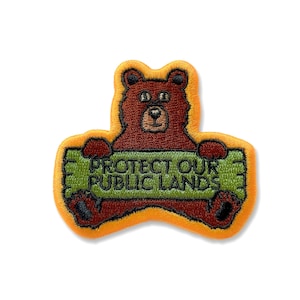 May include: A brown bear holding a green sign that says "Protect Our Public Lands". The bear is smiling and has a brown nose. The sign is outlined in yellow.