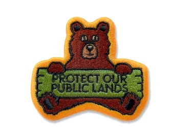 May include: A brown bear holding a green sign that says "Protect Our Public Lands". The bear is smiling and has a brown nose. The sign is outlined in yellow.
