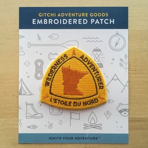 May include: Yellow embroidered patch with the outline of Minnesota, the words "WILDERNESS ADVENTURER" and "L'ETOILE DU NORD" in brown text. The patch is on a white card with the text "GITCHI ADVENTURE GOODS EMBROIDERED PATCH" and "IGNITE YOUR ADVENTURE™".
