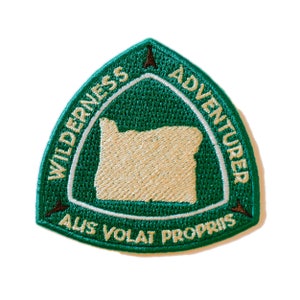 May include: Green embroidered patch with a white outline. The patch is shaped like a triangle with a rounded top. The patch features the text "WILDERNESS ADVENTURER" in a circle around the outline of the state of Oregon. The text "ALIS VOLAT PROPRIS" is below the state outline.