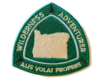 May include: Green embroidered patch with a white outline. The patch is shaped like a triangle with a rounded top. The patch features the text "WILDERNESS ADVENTURER" in a circle around the outline of the state of Oregon. The text "ALIS VOLAT PROPRIS" is below the state outline.