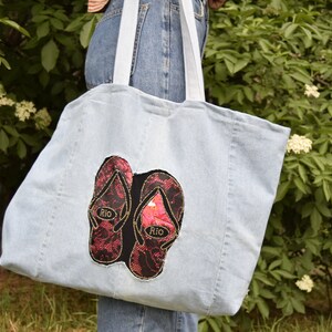 May include: A light blue denim tote bag with a pair of red and black flip flops embroidered on the front. The flip flops have the word "Rio" written on them.