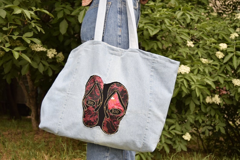 May include: A light blue denim tote bag with a pair of red and black flip flops embroidered on the front. The flip flops have the word "Rio" written on them.