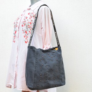 May include: A gray denim tote bag with a black strap and silver studs. The strap has the text "SHINE BRIGHT" repeated on it.
