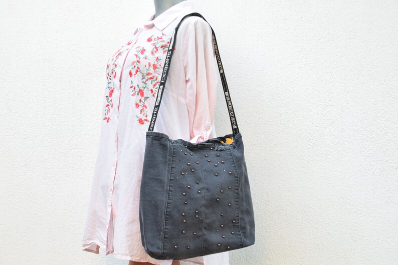 May include: A gray denim tote bag with a black strap and silver studs. The strap has the text "SHINE BRIGHT" repeated on it.