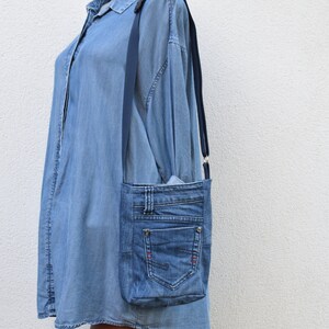 Puede incluir: Bolso bandolera de mezclilla azul claro, hecho de jeans reciclados. Cuenta con un bolsillo frontal, trabillas y una correa ajustable azul oscuro. Detalles de costuras rojas.