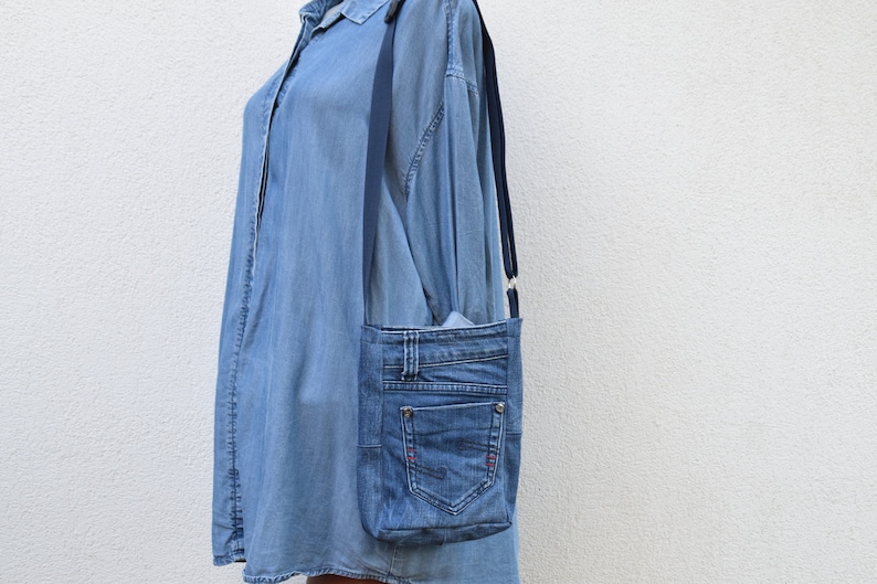 Puede incluir: Bolso bandolera de mezclilla azul claro, hecho de jeans reciclados. Cuenta con un bolsillo frontal, trabillas y una correa ajustable azul oscuro. Detalles de costuras rojas.