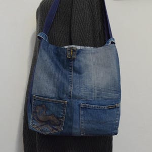 Könnte beinhalten: Eine Jeans-Umhängetasche mit blauem Riemen. Die Tasche besteht aus recycelten Jeans und hat zwei Taschen und ein dekoratives Design auf einer Tasche. Die Tasche hat einen Schnallenverschluss und ein hellblaues Innenfutter.